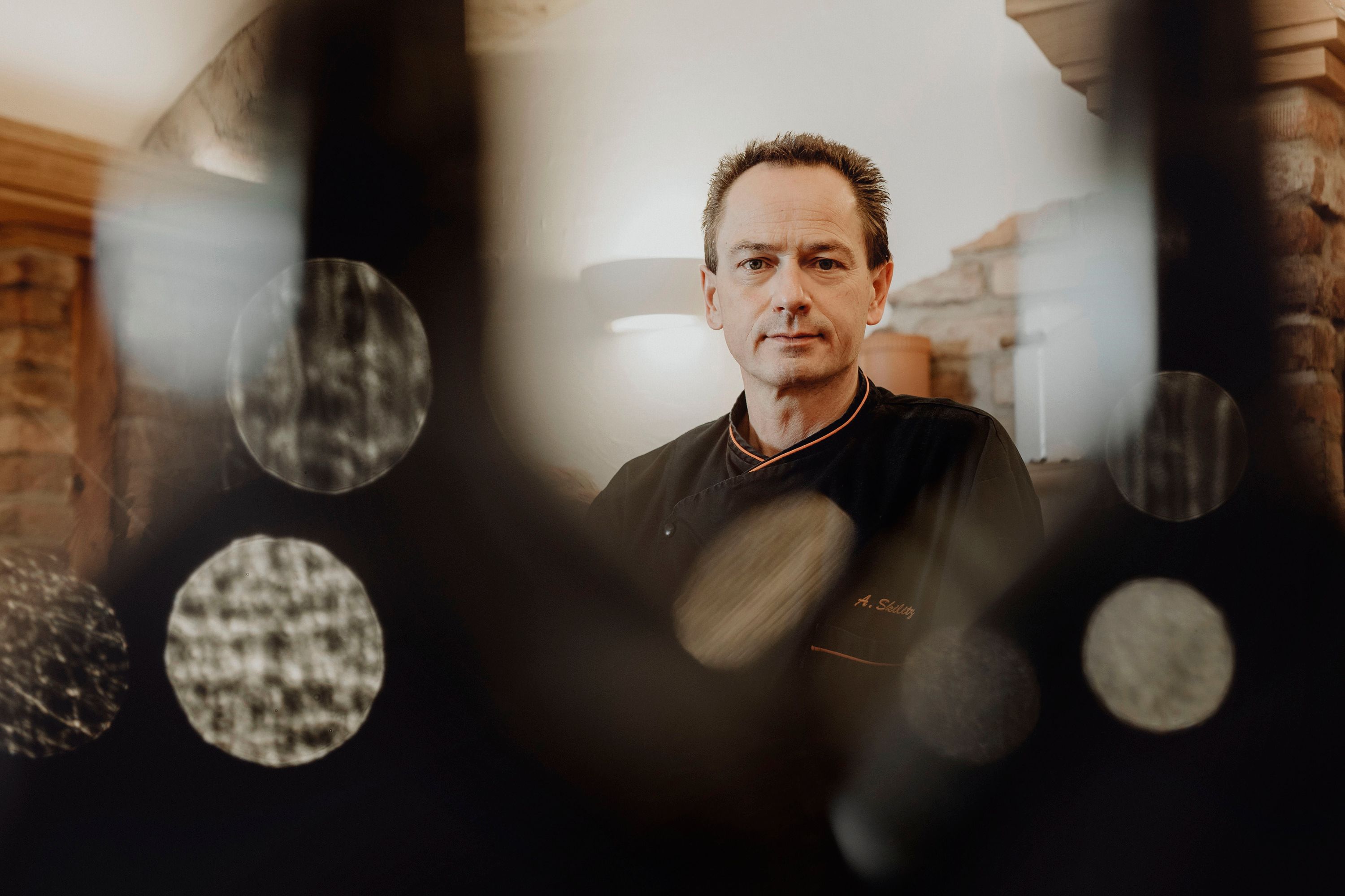 Ein Mann in schwarzer Kochkleidung steht in einem rustikalen Raum, umgeben von unscharfen Lichteffekten.