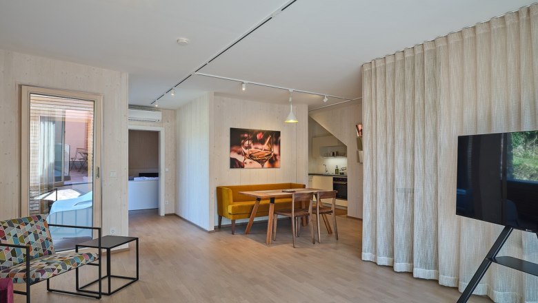 Modernes Wohnzimmer mit Holzboden, Esstisch, gelber Bank, bunter Sessel, gro&szlig;em Fernseher und offener K&uuml;che.