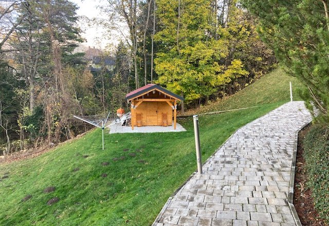 Ein Garten mit einem kleinen Holzhaus, umgeben von Bäumen und einem gepflasterten Weg.