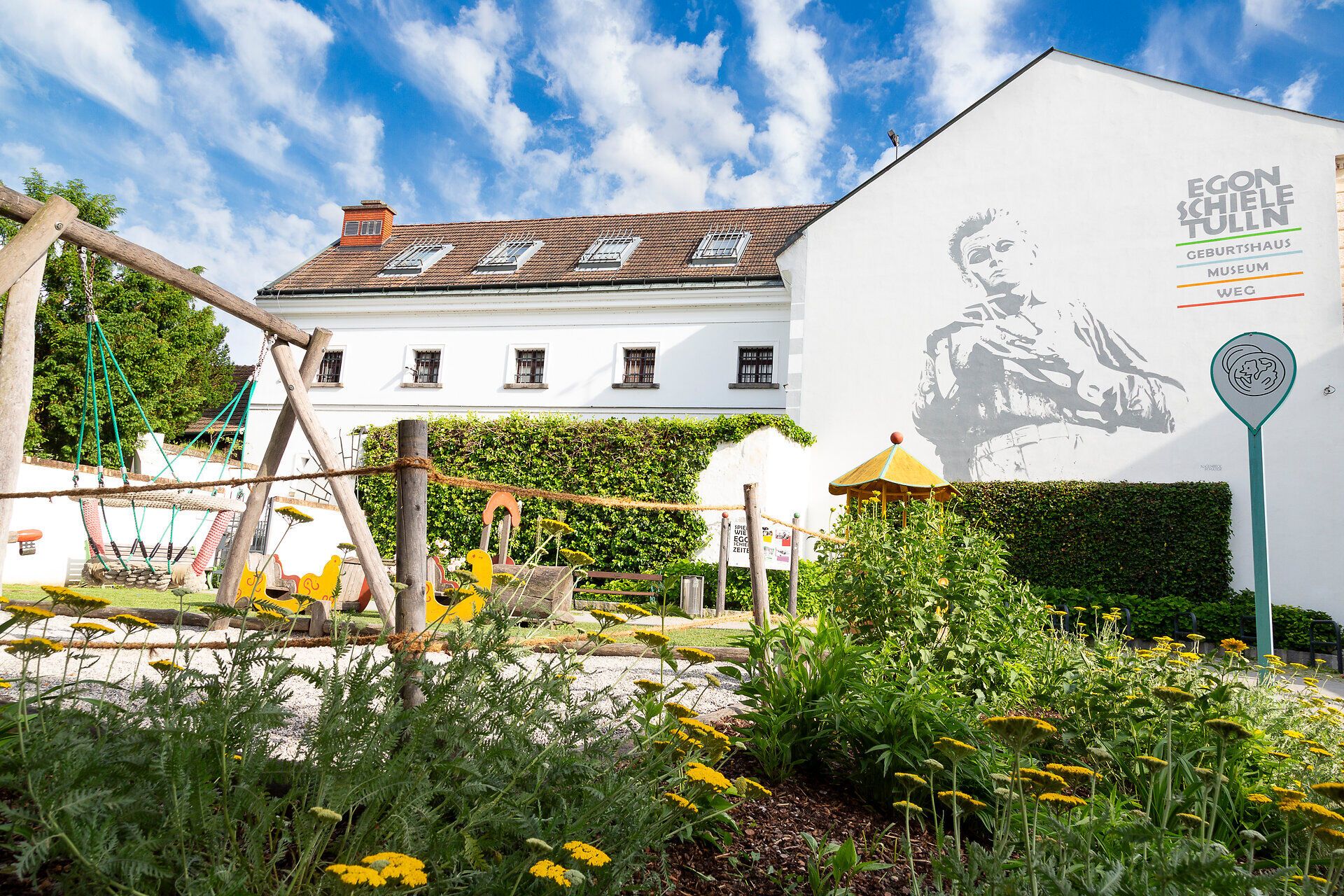 Ein einladender Spielplatz umgeben von blühenden Pflanzen und einer beeindruckenden Wandmalerei, die die Kreativität und den Geist der Kunst feiert. Hier können Familien die frische Luft genießen und die Schönheit der Natur erleben, während die Kinder fröhlich spielen. Ein Ort, der sowohl für Kunstliebhaber als auch für Naturliebhaber ein wahres Paradies ist.
