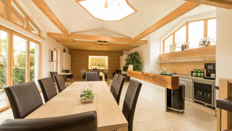 Innenansicht eines modernen Verkostungsraums mit Holztischen und -stühlen, großen Fenstern und einer Bar mit Weinflaschen.