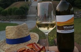 Ein Glas Weißwein und eine Flasche vor einem Turm, mit Käse, Schinken und Brot auf einem Holzbrett.