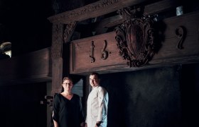 Zwei Personen stehen vor einem alten Holzbalken mit Schnitzereien und der Jahreszahl 1692.