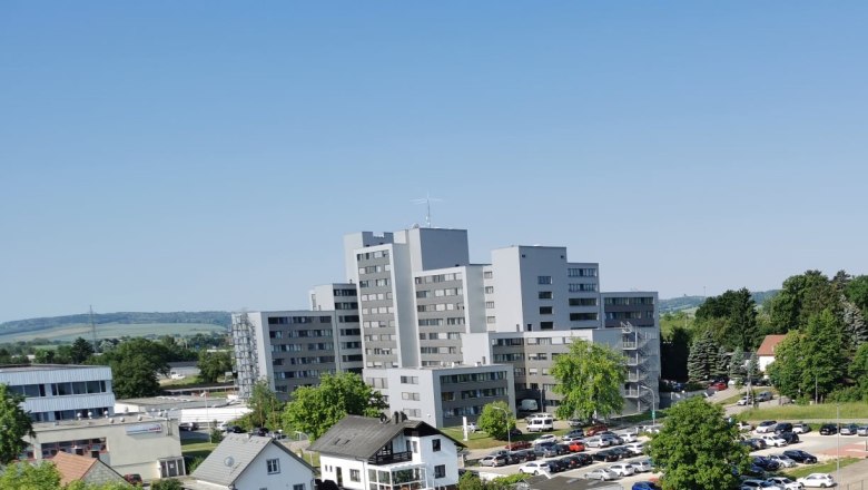 Mehrstöckiges graues Gebäude mit umliegenden Bäumen und Autos auf einem Parkplatz, blauer Himmel im Hintergrund.