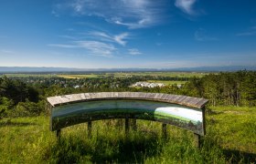 Panoramablick von einem Aussichtspunkt mit Infotafel, im Hintergrund eine weite Landschaft und blauer Himmel.
