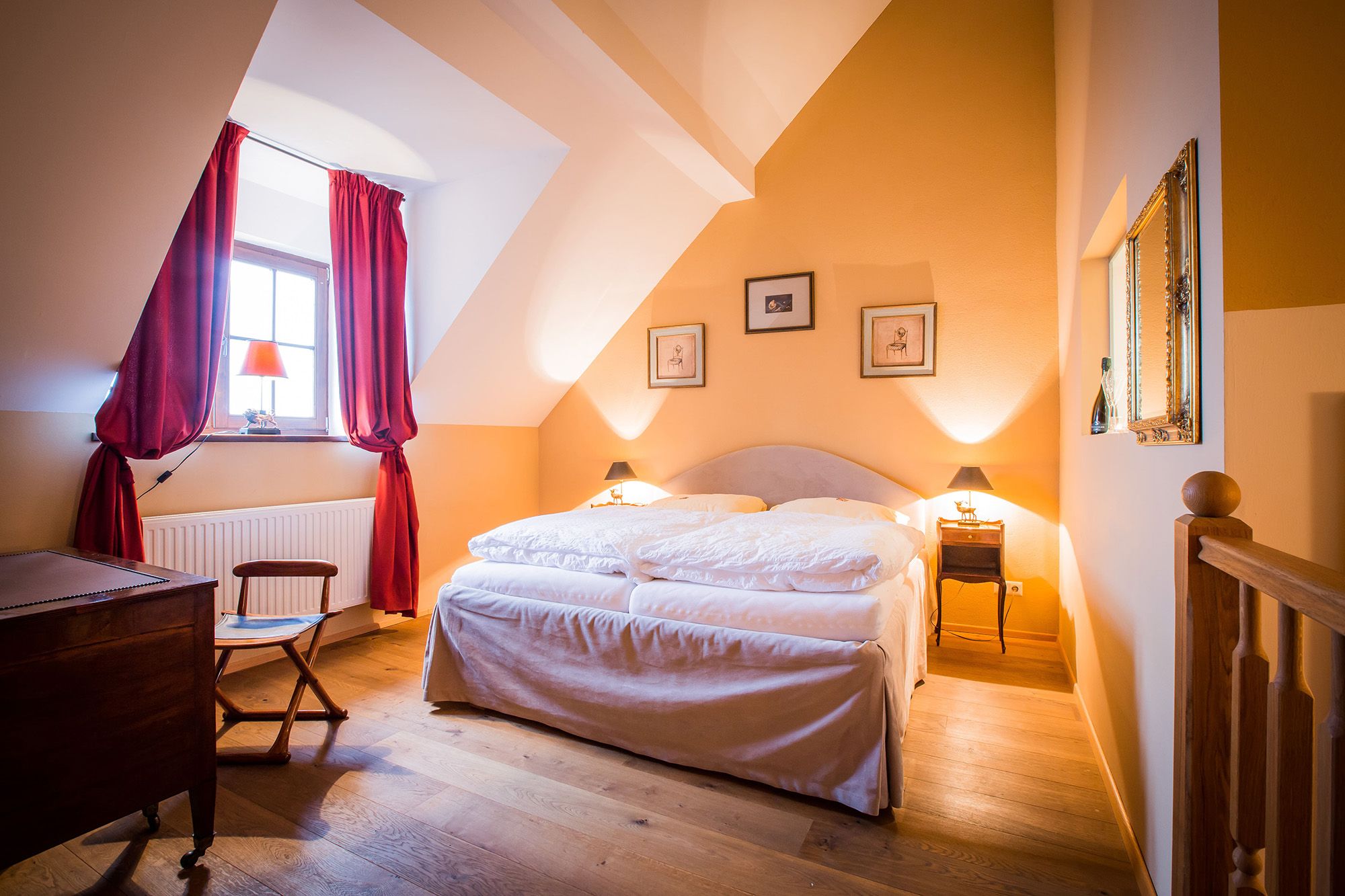 Ein gemütliches Doppelzimmer mit einem großen Bett, roten Vorhängen und warmen Farben. An der Wand hängen Bilder, und ein Fenster lässt Tageslicht herein.