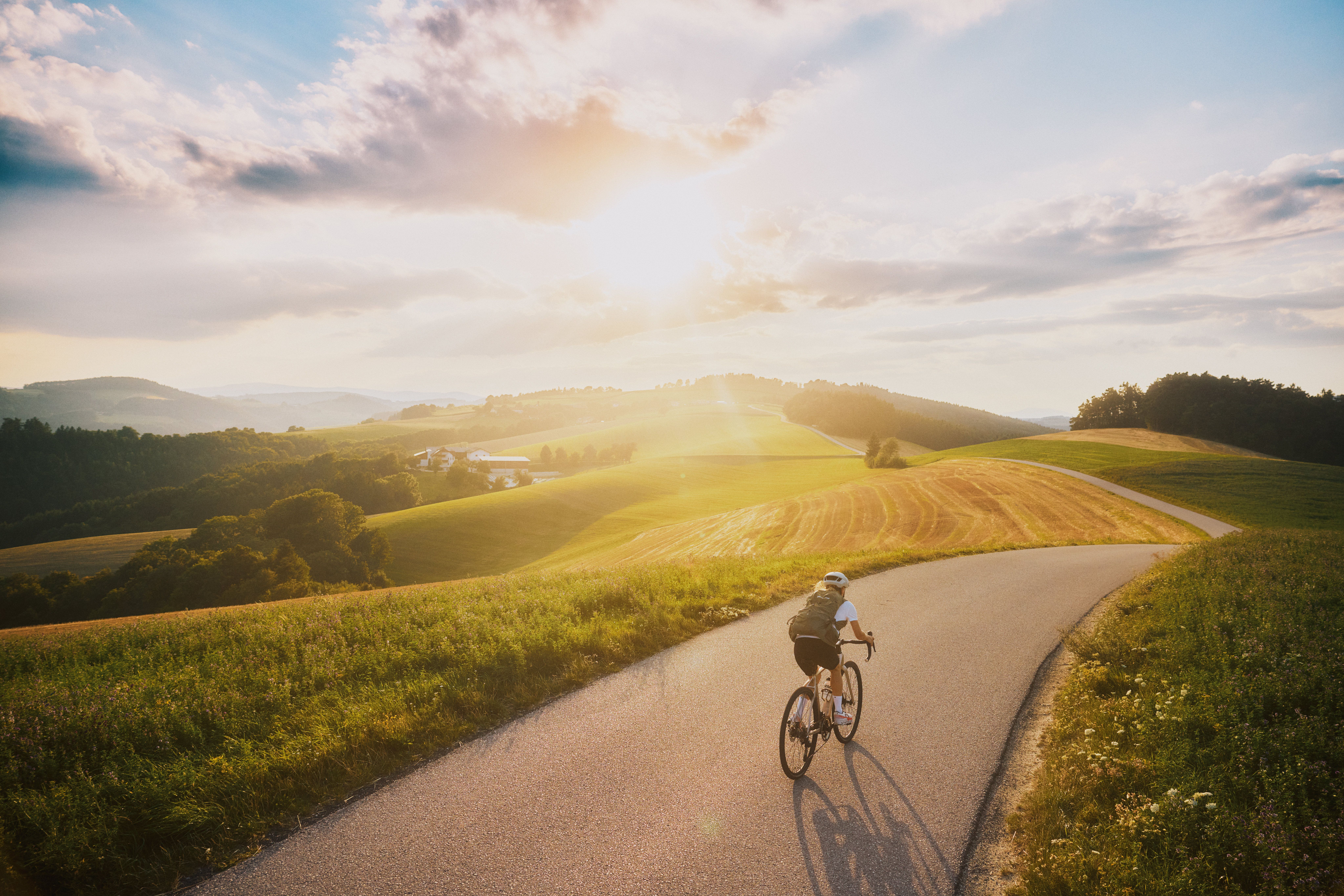 Eine Radfahrerin fährt auf einer kurvigen Landstraße durch eine hügelige Landschaft bei Sonnenuntergang.