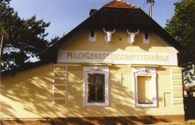 Gelbes Gebäude mit der Aufschrift 'Milchgenossenschaftsgebäude' und zwei Stierköpfen an der Fassade.