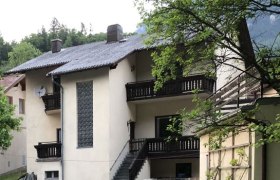 Ein zweist&ouml;ckiges Haus mit Balkon und Treppe, umgeben von einem gr&uuml;nen Garten mit Blumen.