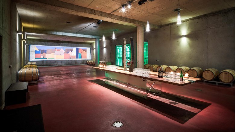 Ein moderner Weinkeller mit Holzf&auml;ssern und einem langen Tisch mit Gl&auml;sern und Flaschen.