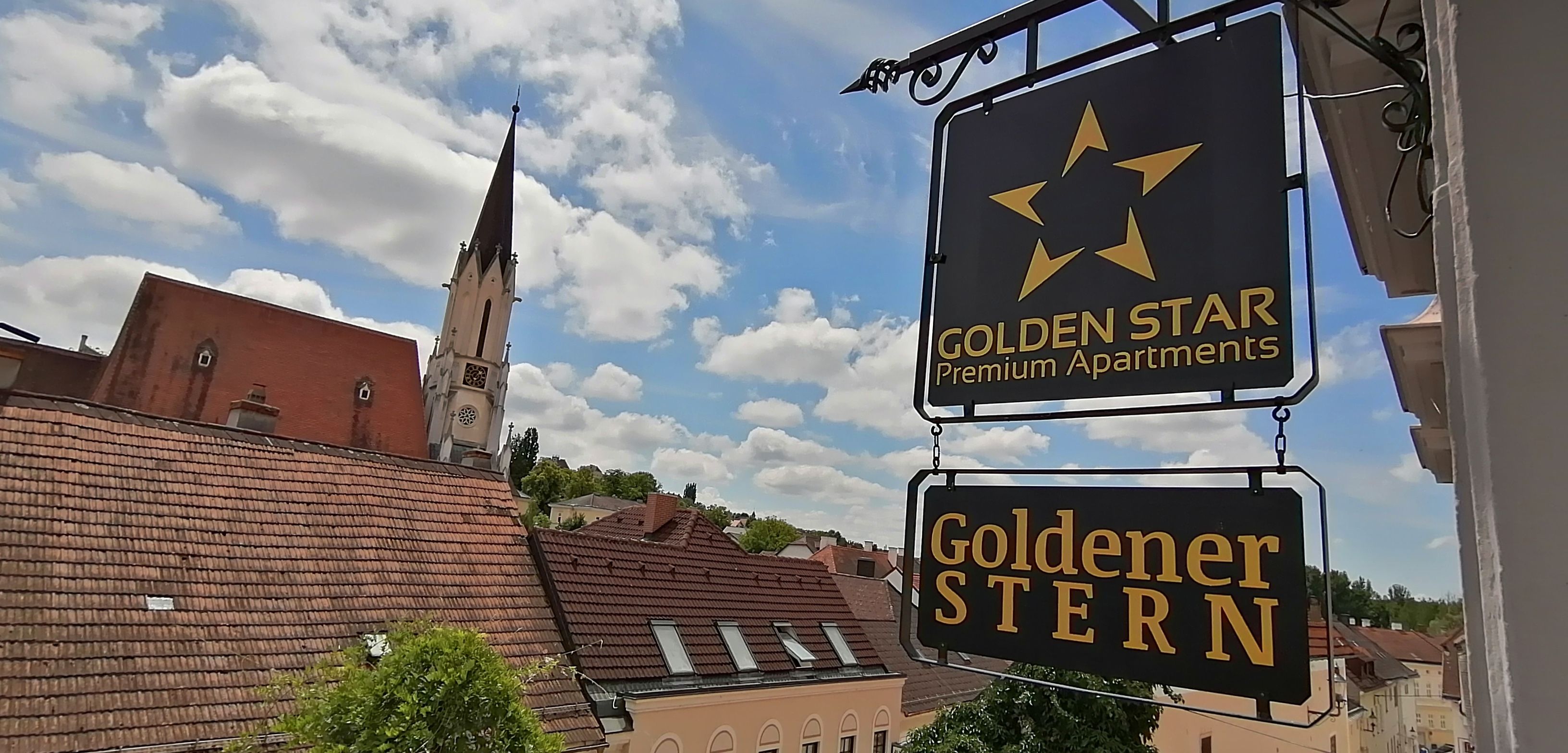 Blick auf ein Schild mit der Aufschrift 'Golden Star Premium Apartments' und 'Goldener Stern' vor einem Kirchturm und Dächern.