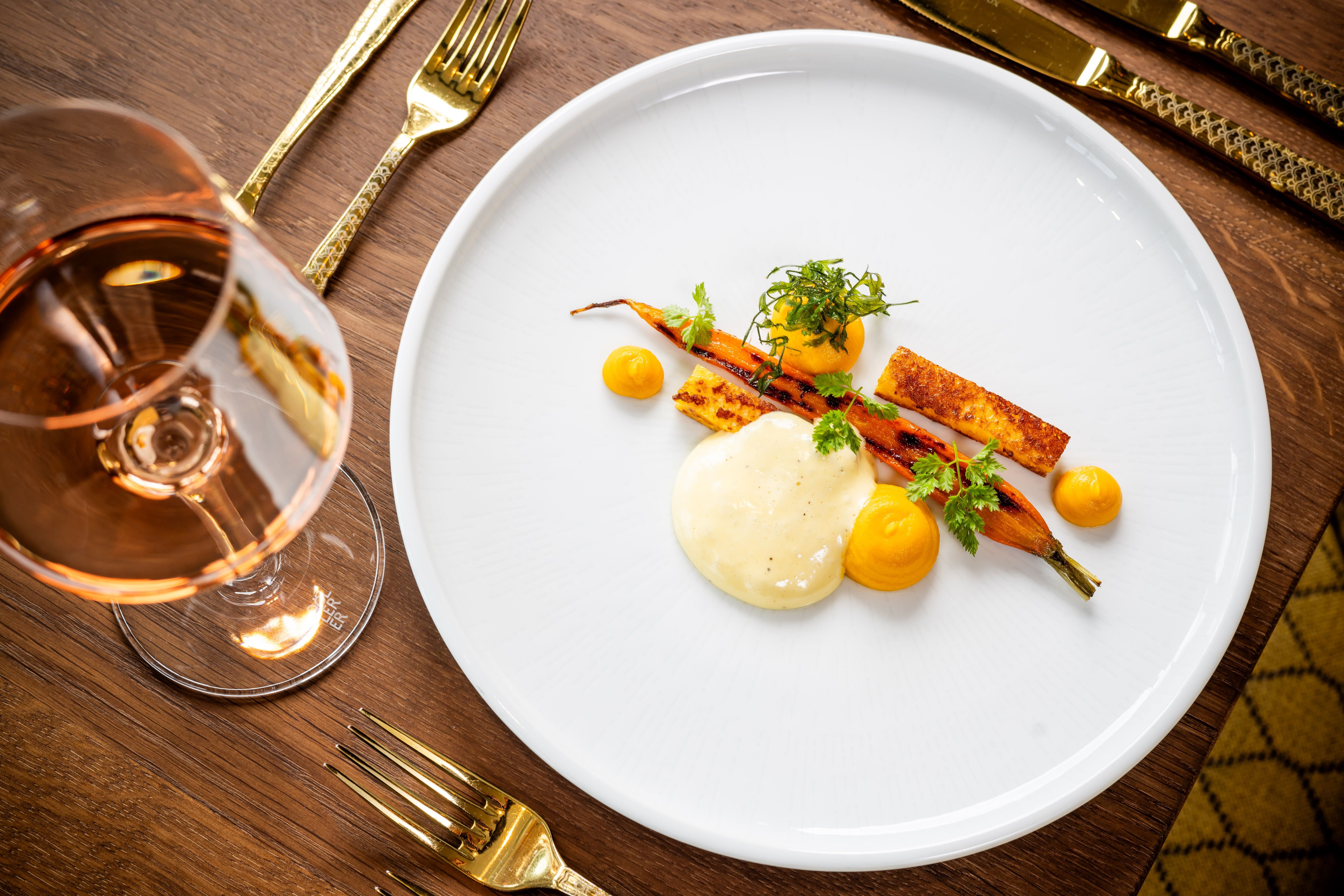 Ein elegantes Gericht mit Karotten, Polenta, Heu und Kerbel auf einem weißen Teller, daneben ein Glas Roséwein auf einem Holztisch.