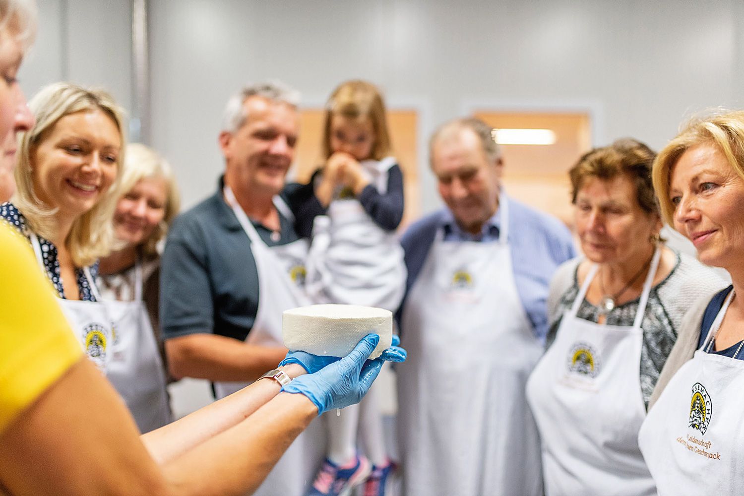 Eine Gruppe von Menschen betrachtet einen frisch hergestellten Käse, der von einer Person mit blauen Handschuhen gehalten wird.