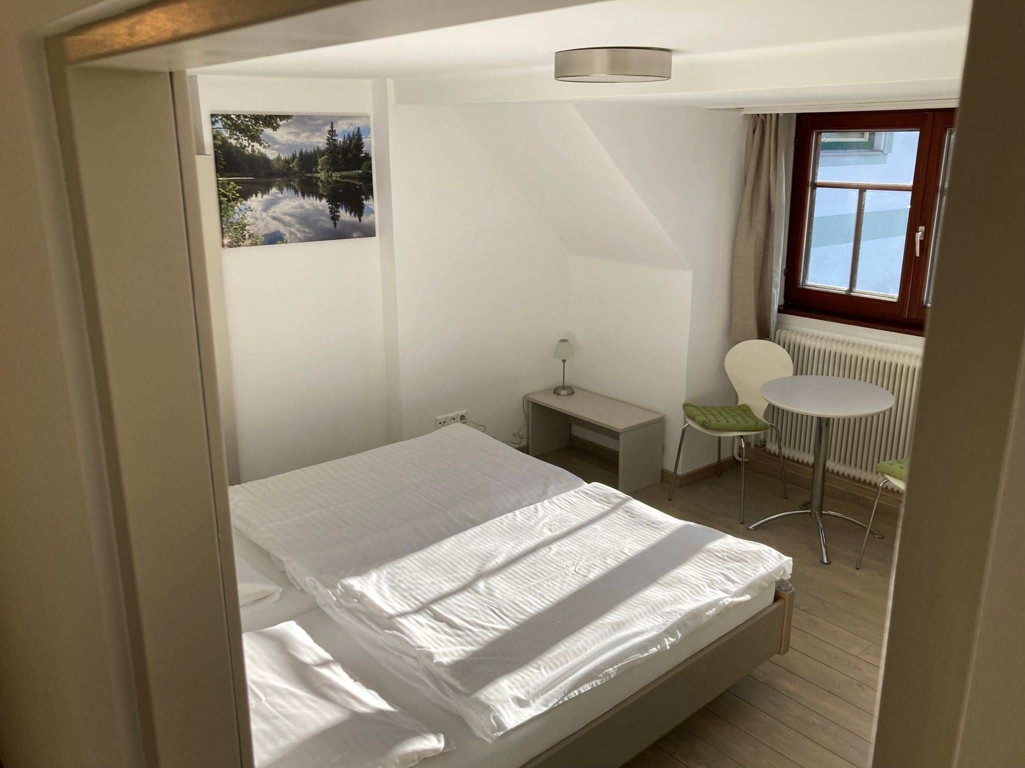 Ein schlicht eingerichtetes Zimmer mit einem Bett, einem kleinen Tisch und zwei Stühlen. Ein Fenster lässt Tageslicht herein, und ein Bild hängt an der Wand.
