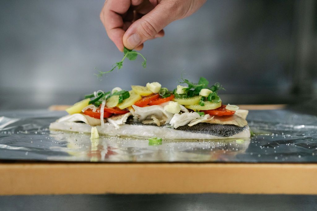 Ein Fischfilet mit Gemüse und Kräutern auf Alufolie, eine Hand fügt Garnitur hinzu.