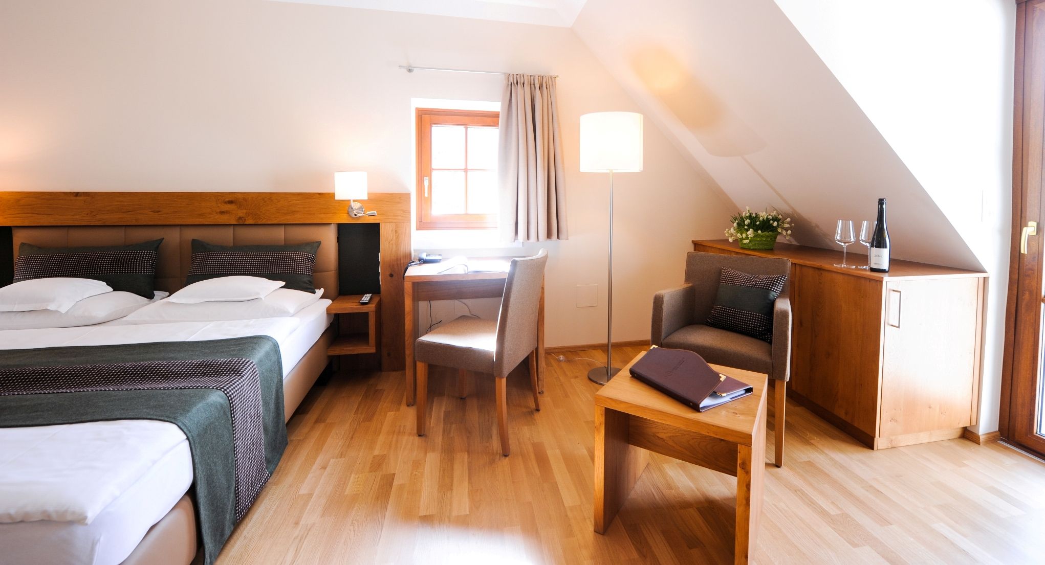 Gemütliches Hotelzimmer mit Doppelbett, Schreibtisch, Sessel und Flasche Wein auf einem Schrank.