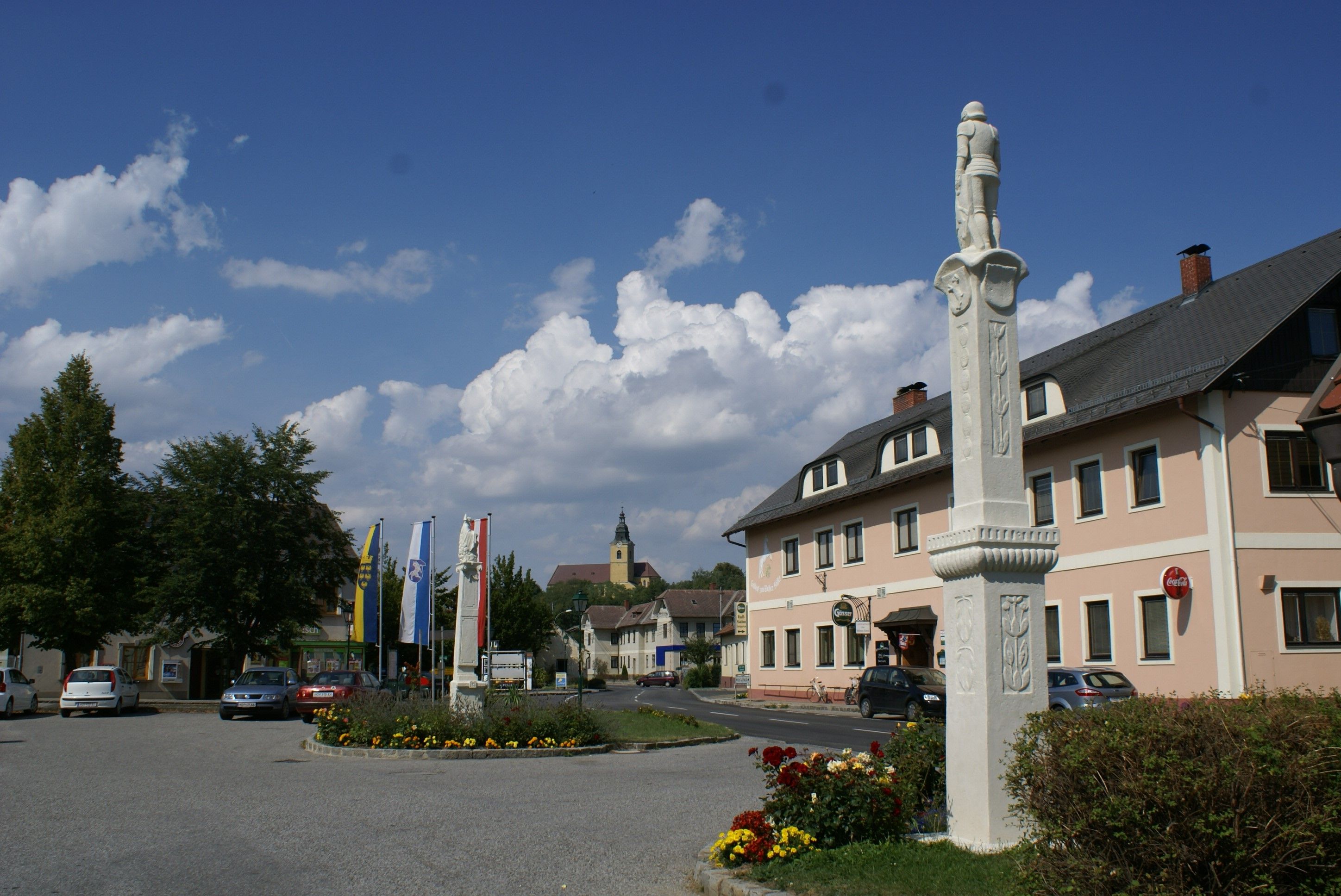 Ein Platz in Weitersfeld mit einer Statue, mehreren Flaggen und einem Gebäude im Hintergrund.