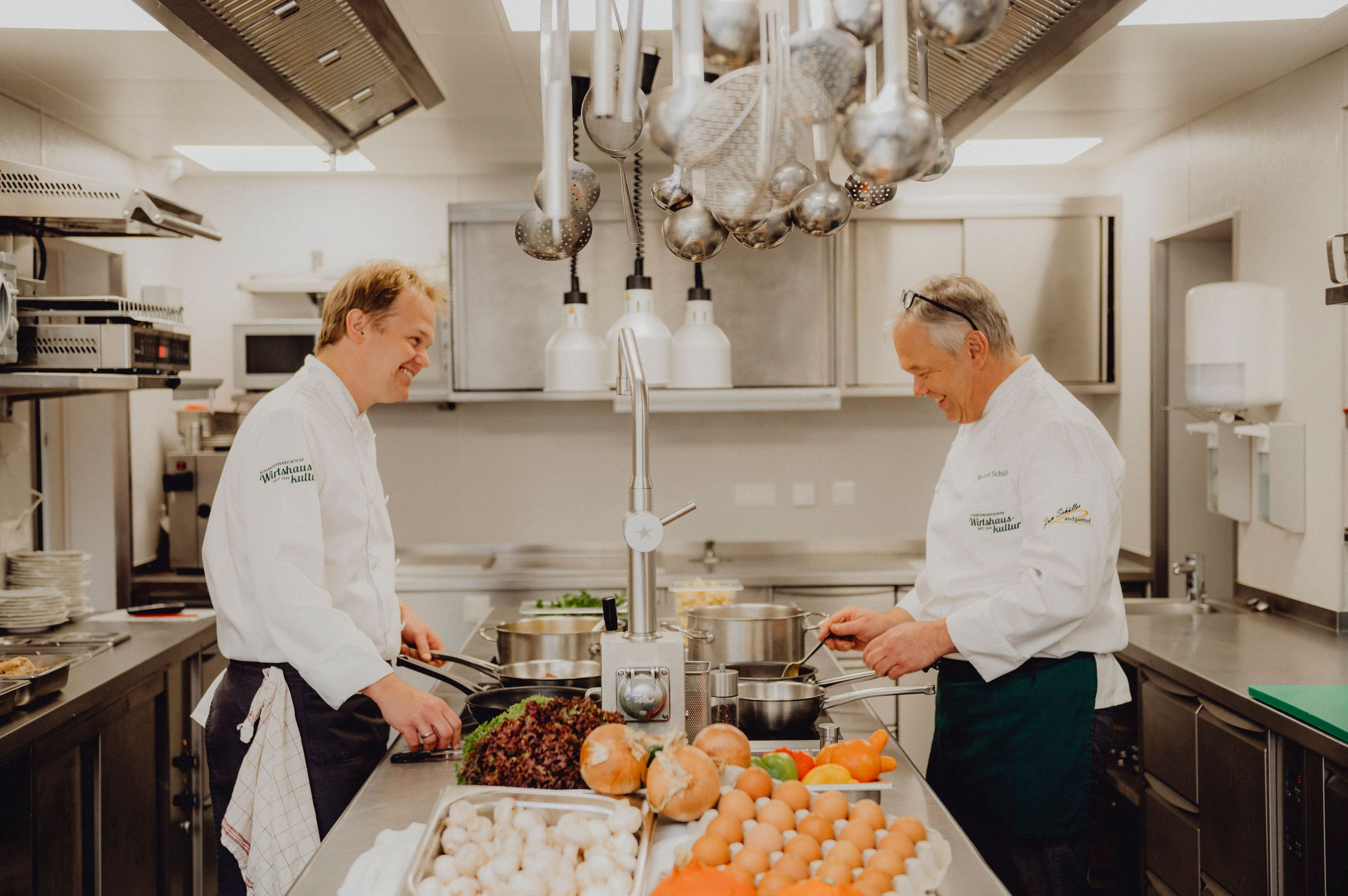 Zwei Köche in einer professionellen Küche, die gemeinsam kochen. Auf dem Tisch liegen Gemüse und Eier.
