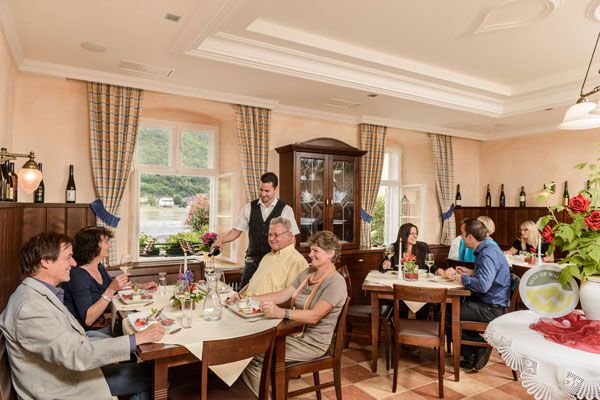 Ein gemütliches Restaurant mit Gästen an Tischen, die speisen. Ein Kellner bedient die Gäste. Fenster mit Blick auf eine grüne Landschaft.