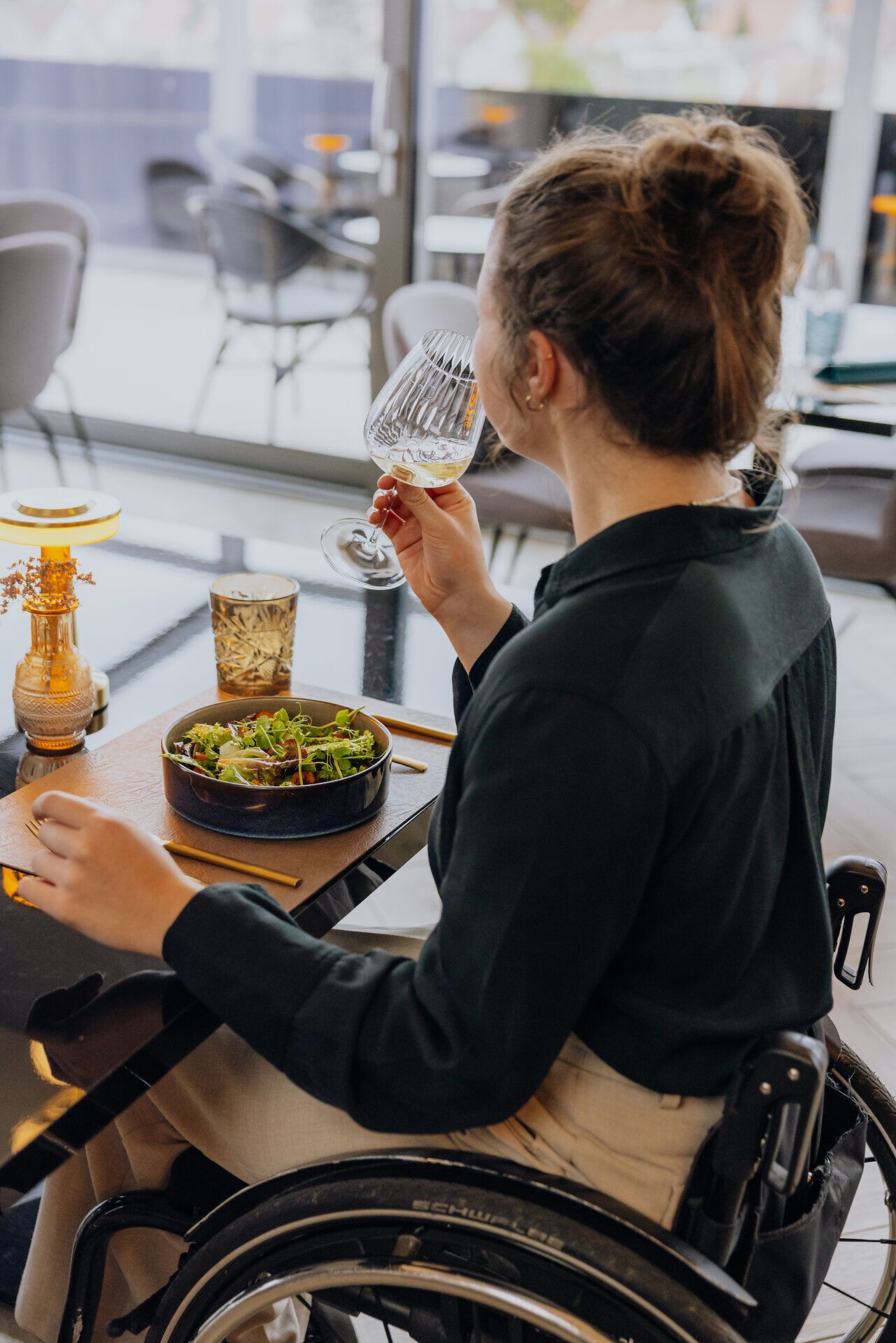 Eine Frau im Rollstuhl sitzt in einem Restaurant an einem Tisch und kostet an einem Glas Weißwein. Vor ihr auf dem Tisch steht ein herbstlicher Salat.