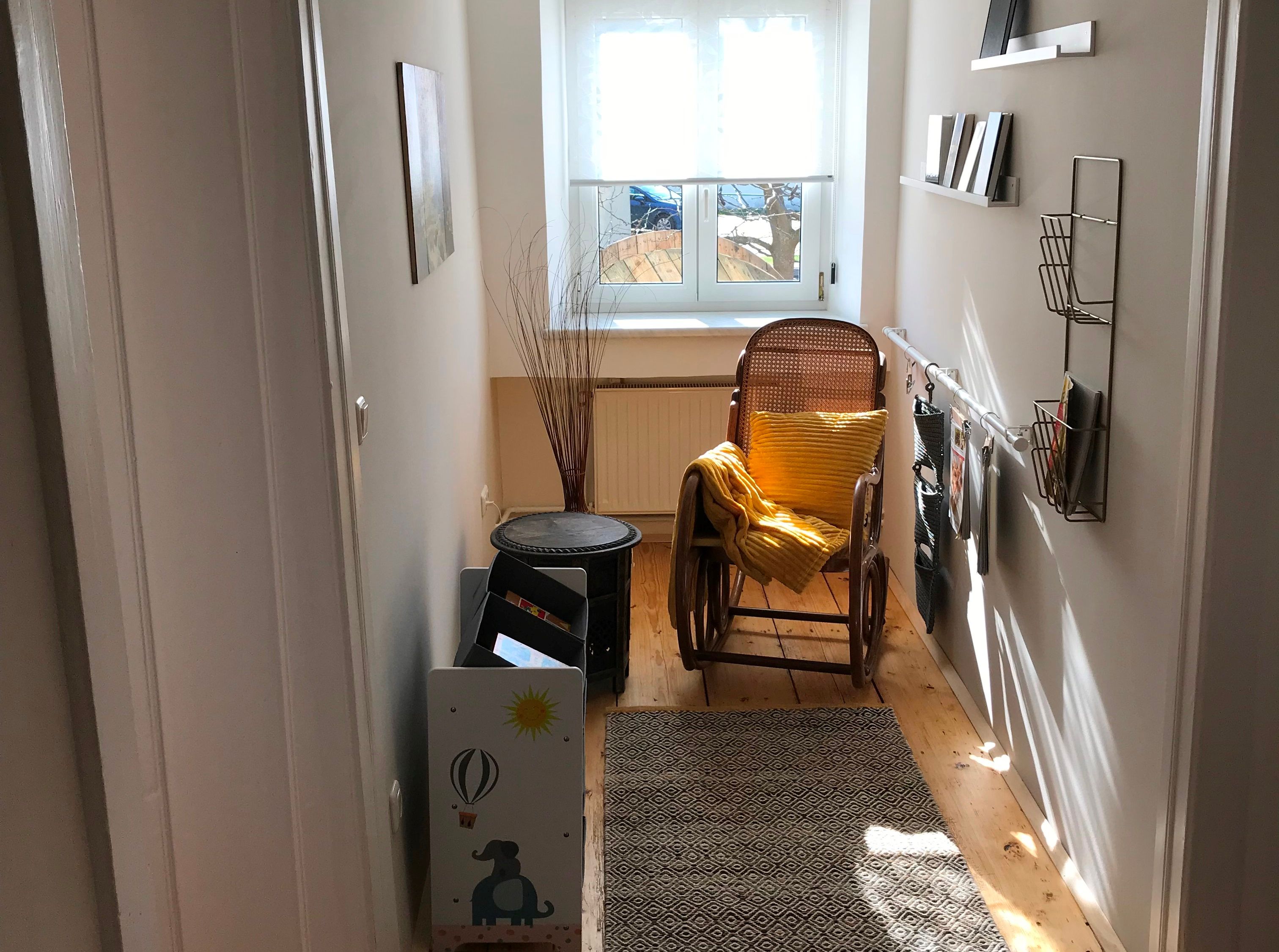 Ein schmaler Eingangsbereich mit Schaukelstuhl, gelbem Kissen, Teppich und Regalen an der Wand. Ein Fenster am Ende des Flurs.