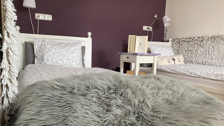 Zweibettzimmer mit lila Wand, zwei Betten, Nachttisch mit Buch und flauschiger Decke.