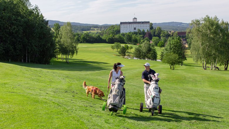 Zwei Golfer mit Hund auf gr&uuml;nem Golfplatz, Schloss im Hintergrund.