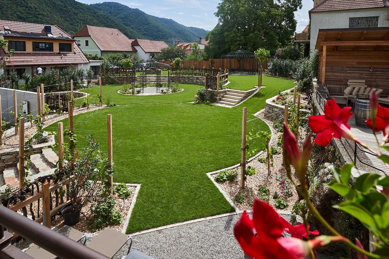 Ein gepflegter Hotelgarten mit grünem Rasen, Blumenbeeten und einem Pavillon, umgeben von Häusern und Hügeln im Hintergrund.