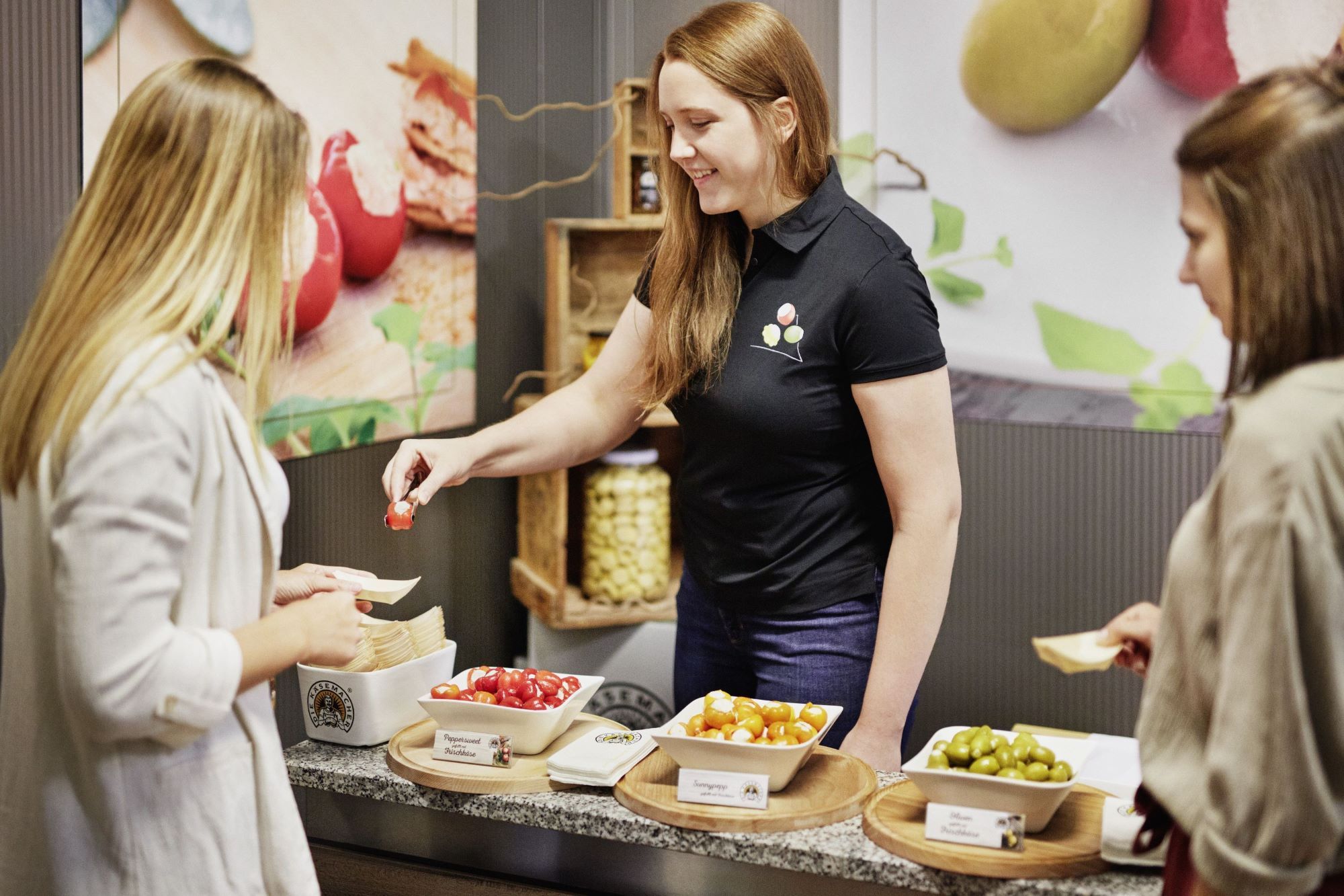 Eine Frau bietet zwei anderen Frauen verschiedene gefüllte Paprikas und Oliven zur Verkostung an.