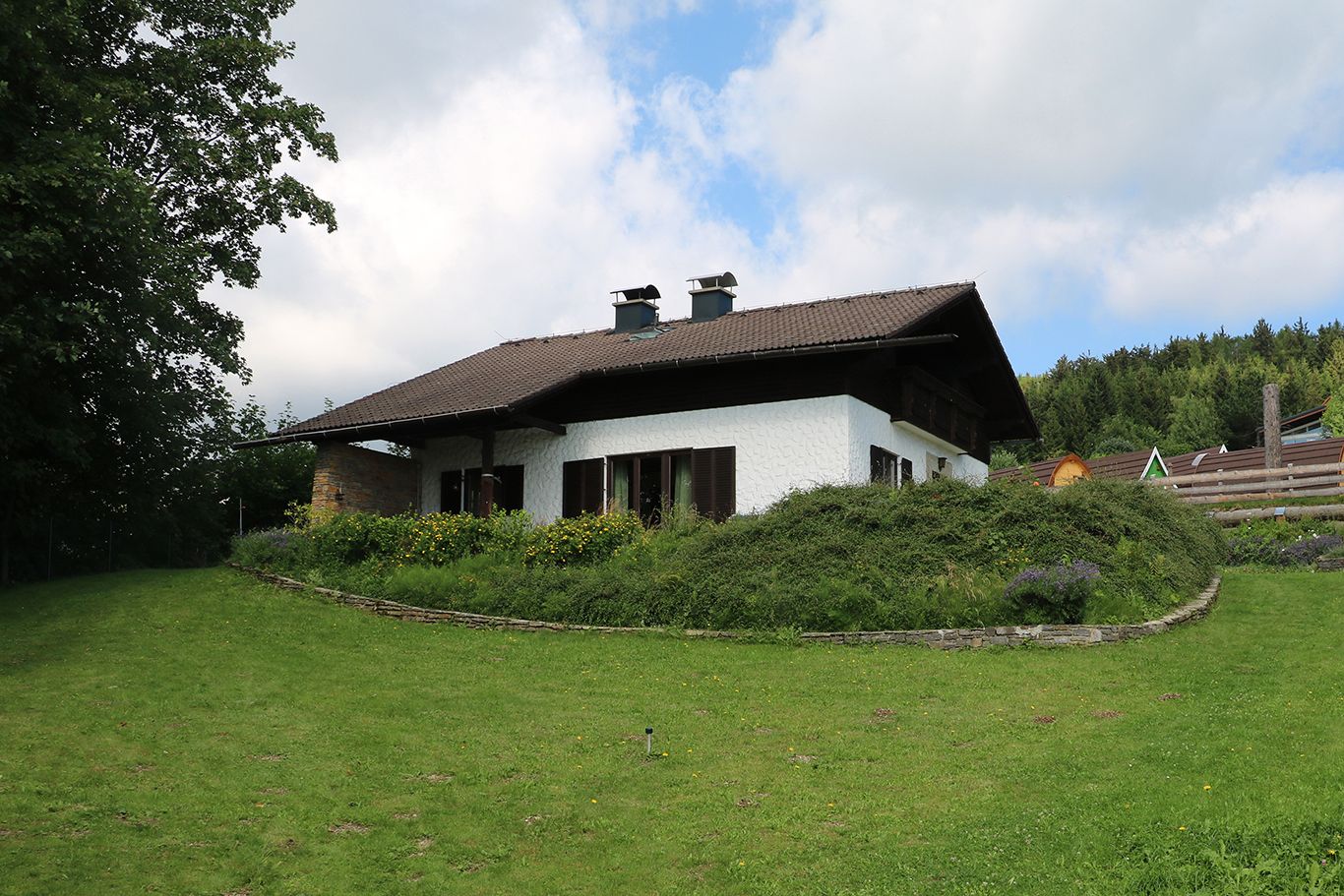 Ein weißes Haus mit braunem Dach in einem grünen Garten mit Bäumen und Blumen.