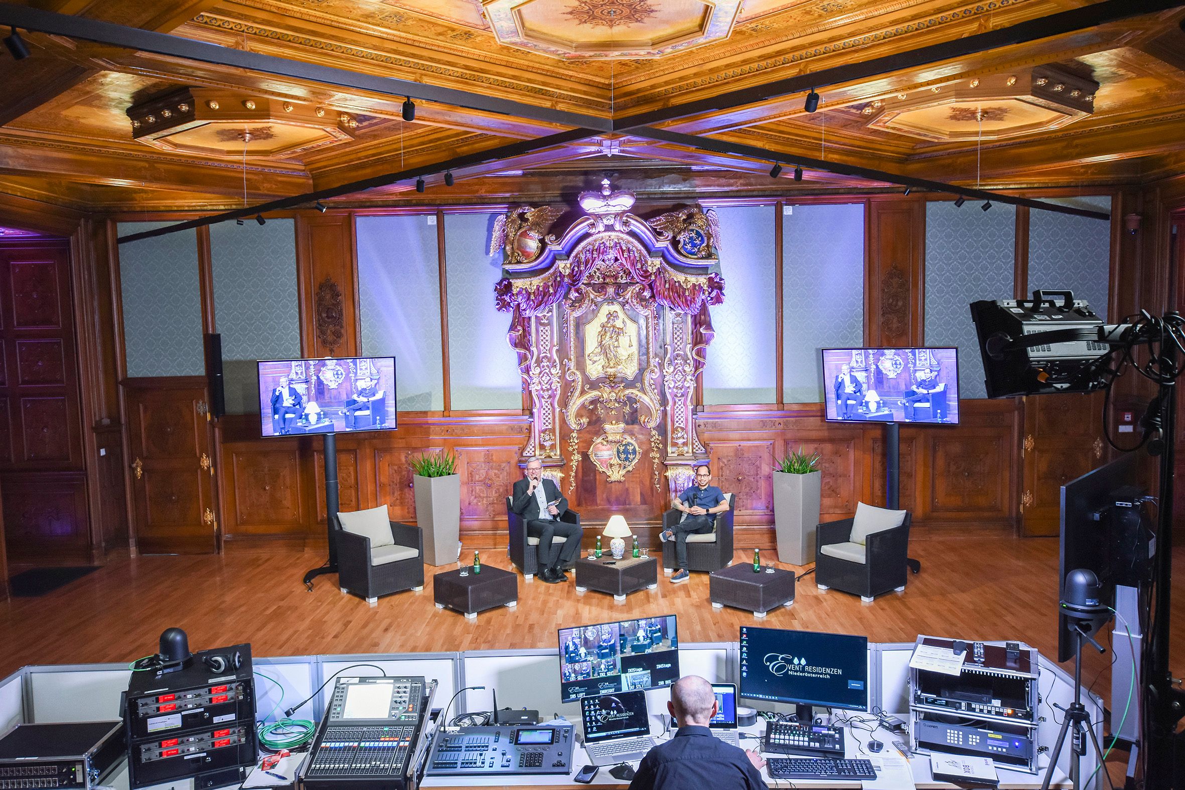 Ein opulenter Raum mit Holzvertäfelung und einem aufwendigen Altar. Zwei Personen sitzen auf einer Bühne, umgeben von Technik und Bildschirmen.