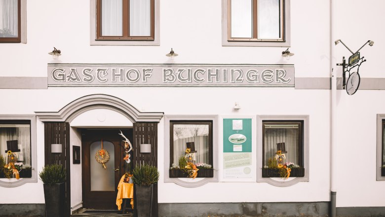 Fassade eines Gasthofs mit der Aufschrift 'Gasthof Buchinger'. Dekorationen und Pflanzen schm&uuml;cken den Eingang.