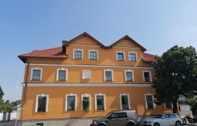 Gelbes Gebäude mit roten Dachziegeln und mehreren Fenstern, davor geparkte Autos und ein Baum.