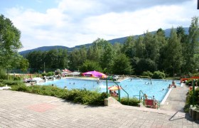 Freibad mit Schwimmern und Sonnenschirmen, umgeben von B&auml;umen und H&uuml;geln.