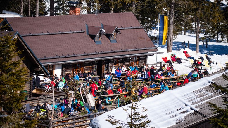 Menschen sitzen vor einer Berghütte im Schnee, umgeben von Bäumen.