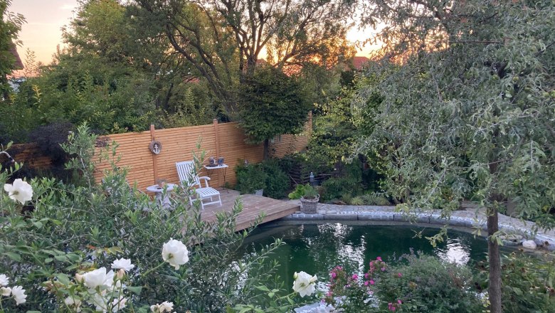 Ein idyllischer Garten mit Teich, Holzterrasse und wei&szlig;en Gartenst&uuml;hlen, umgeben von &uuml;ppiger Vegetation und bl&uuml;henden Blumen.