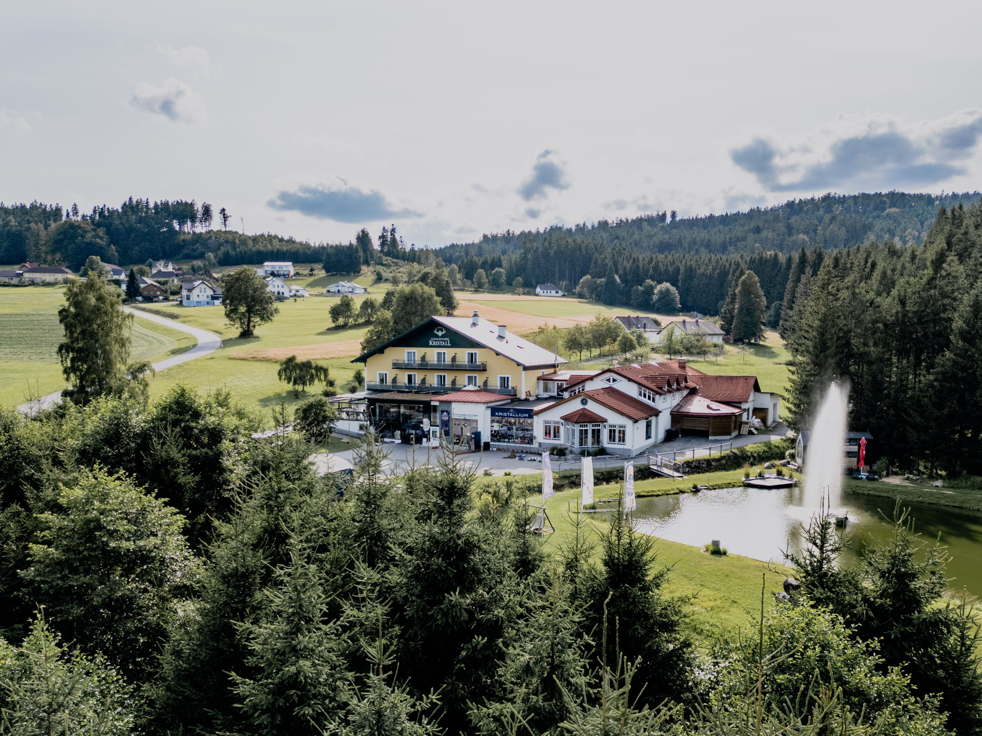 Landschaft mit Gebäuden, Teich und Wald im Hintergrund.