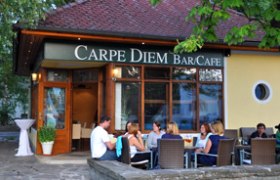 Außenansicht eines Cafés mit dem Namen Carpe Diem, Menschen sitzen an Tischen im Freien.