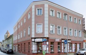 Dreistöckiges Hotelgebäude an einer Straßenecke mit Autos und Geschäften.