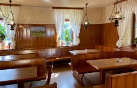 Gem&uuml;tliche Gaststube mit Holzm&ouml;beln, wei&szlig;en Vorh&auml;ngen und Pflanzen auf der Fensterbank.