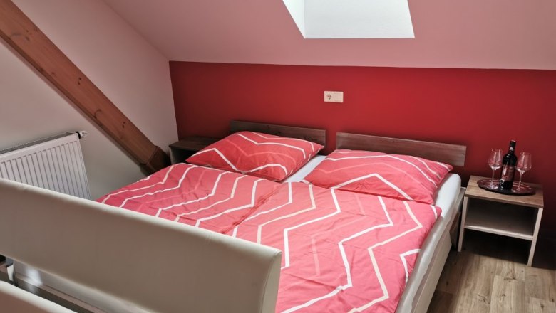Schlafzimmer mit roter Wand, Doppelbett mit roter Bettw&auml;sche, Dachfenster und Nachttisch mit Weinflasche und Gl&auml;sern.