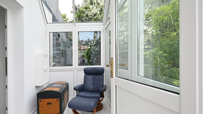 Ein heller Vorraum mit einem blauen Sessel und einer dekorativen Truhe, umgeben von gro&szlig;en Fenstern mit Blick auf gr&uuml;ne Pflanzen.