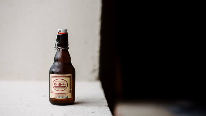 Eine Flasche RaxBräu Helles Bier auf einer weißen Oberfläche vor einem dunklen Hintergrund.