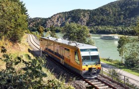 Ein gelber Zug der Wachaubahn f&auml;hrt entlang eines Flusses, im Hintergrund ein Kreuzfahrtschiff und bewaldete H&uuml;gel.