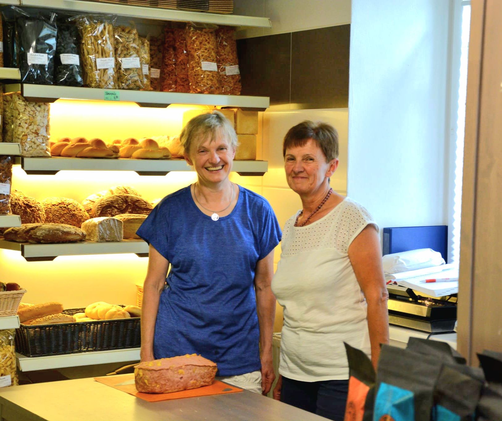 Zwei Frauen in einer Bäckerei vor Regalen mit Brot und Gebäck.