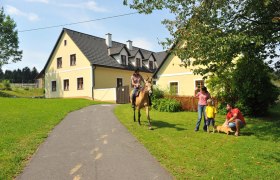 Ein Reiter auf einem Pferd nähert sich einer Familie vor einem gelben Haus auf einem Bauernhof.