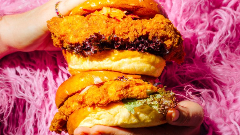 Zwei große, frittierte Chicken-Burger werden von Händen vor einem pinken, flauschigen Hintergrund gehalten.