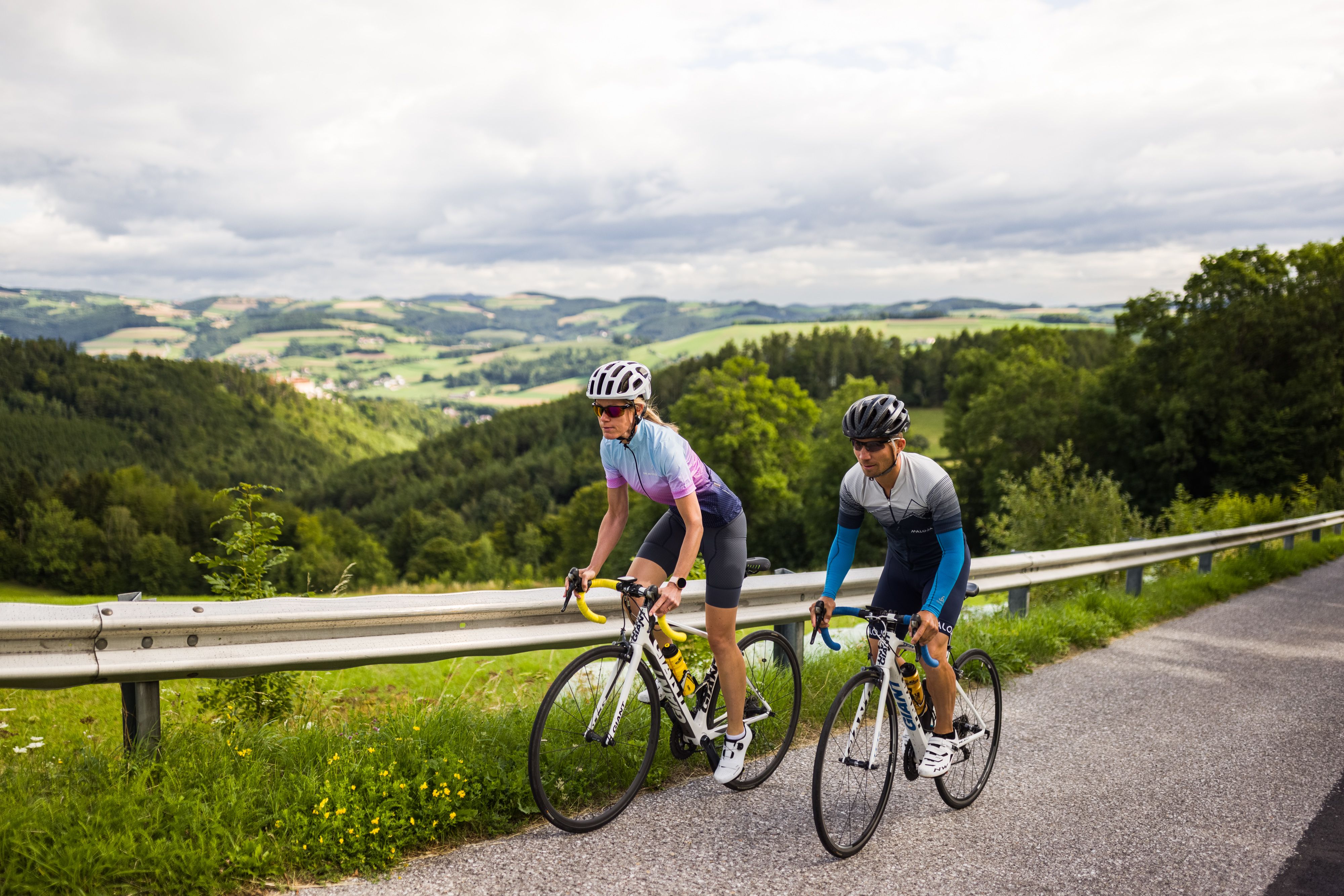 Zwei Radfahrende fahren auf einer Straße in einer hügeligen Landschaft.