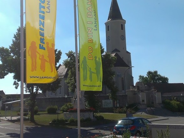 Zwei bunte Fahnen mit der Aufschrift 'Freizeitgemeinde Langau' und 'Do bin i daham' vor einer Kirche.