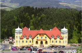 Gelbes Hotel mit roten D&auml;chern in einer gr&uuml;nen, h&uuml;geligen Landschaft.